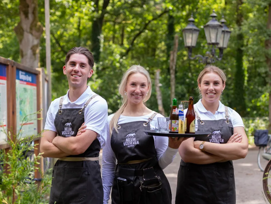 Three staff members of Boshuys Nieuw Vosseven wearing aprons, standing in the forest with drinks on a tray, with a walking path and lanterns in the background