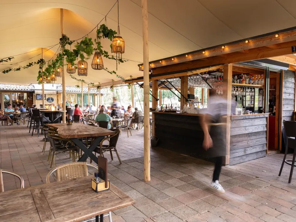 Covered terrace of Boshuys Nieuw Vosseven with wooden tables, chairs, a bar and hanging plants, where guests are seated and a staff member walks by