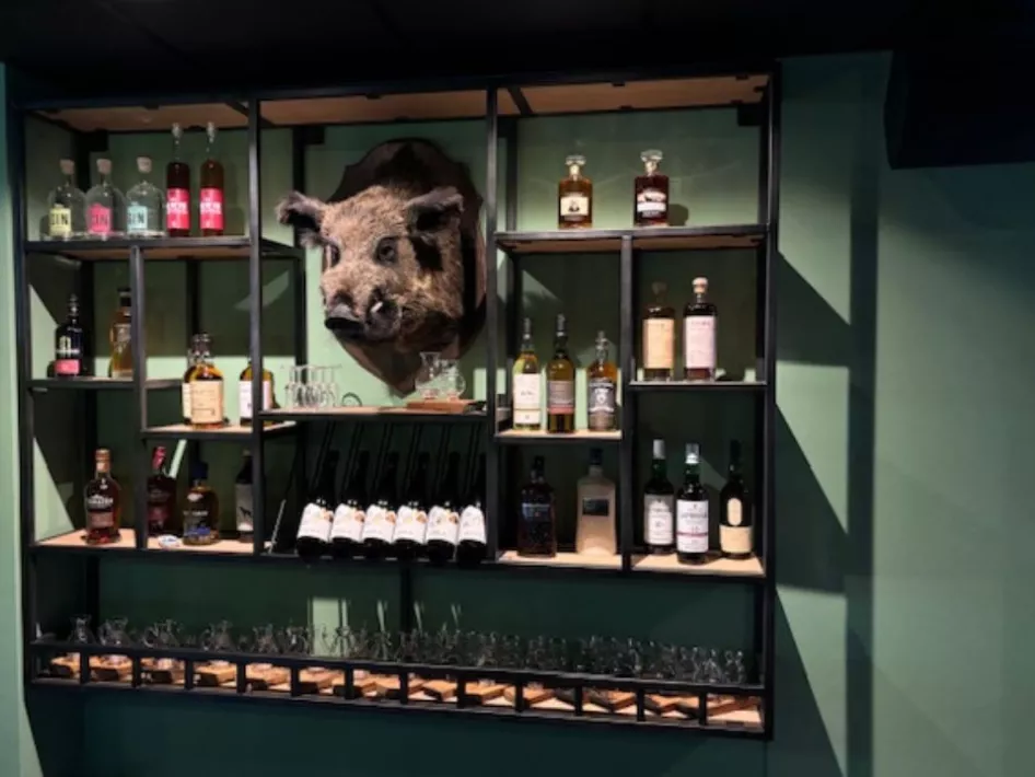  Een wandrek met een uitgebreide selectie whisky’s, gin en bieren, met als opvallend detail een opgezette zwijnenkop aan de muur.