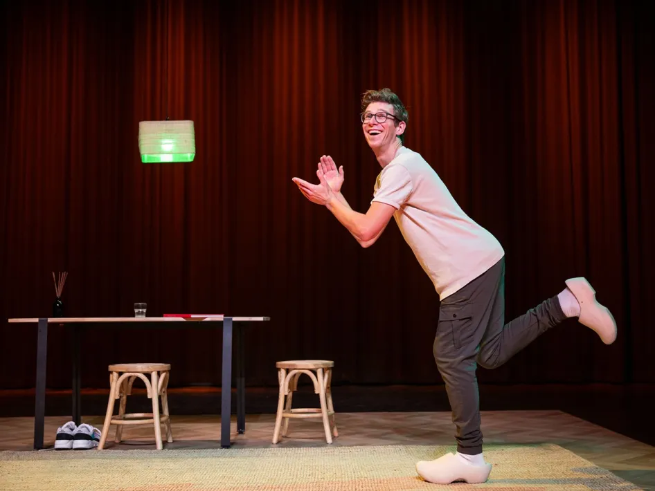 Remy Evers op het podium tijdens cabaretvoorstelling, in een speelse houding met opgetild been, tegen een rode theaterachtergrond met tafel, krukken en lamp.