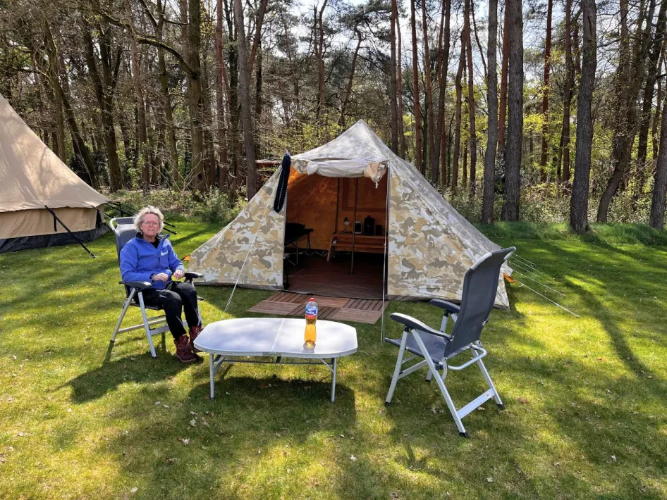 Eine Person sitzt entspannt in einem Campingstuhl vor einem tarnfarbenen Zelt auf einer von Bäumen und anderen Zelten umgebenen Wiese.