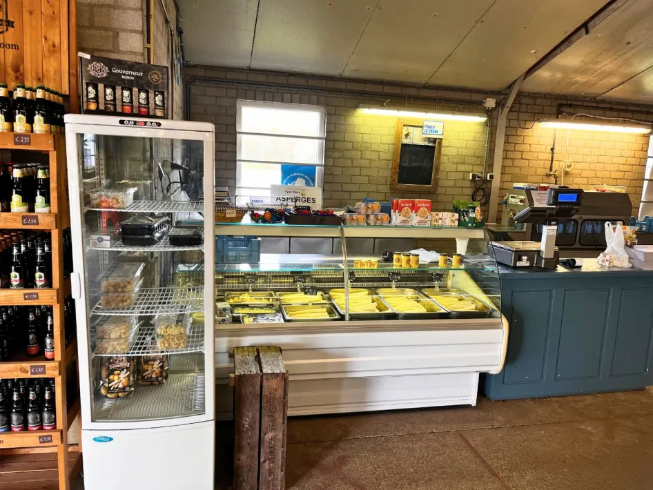 Interior of a farm store with asparagus in the refrigerated counter, local produce in a refrigerator and a rack of local beverages.