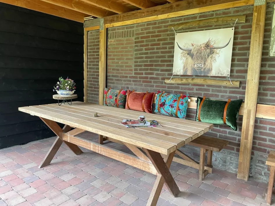 Gezellige overdekte zithoek met een houten tafel, kleurrijke kussens en een decoratief doek met een Schotse hooglander.