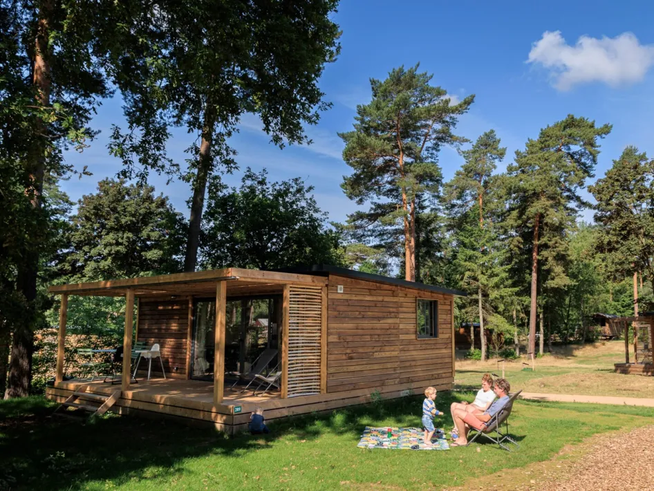 Family with child playing on the grass in front of a wooden holiday home among the trees at Huttopia de Meinweg