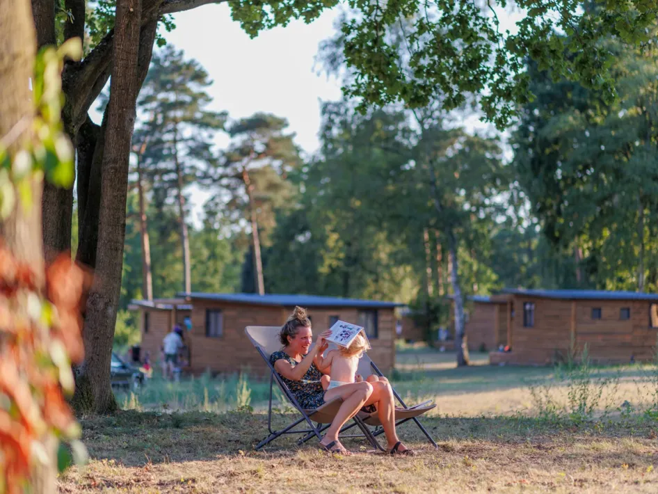 Woman and child relaxing together on a lounge chair in the shade, with wooden holiday homes in the background