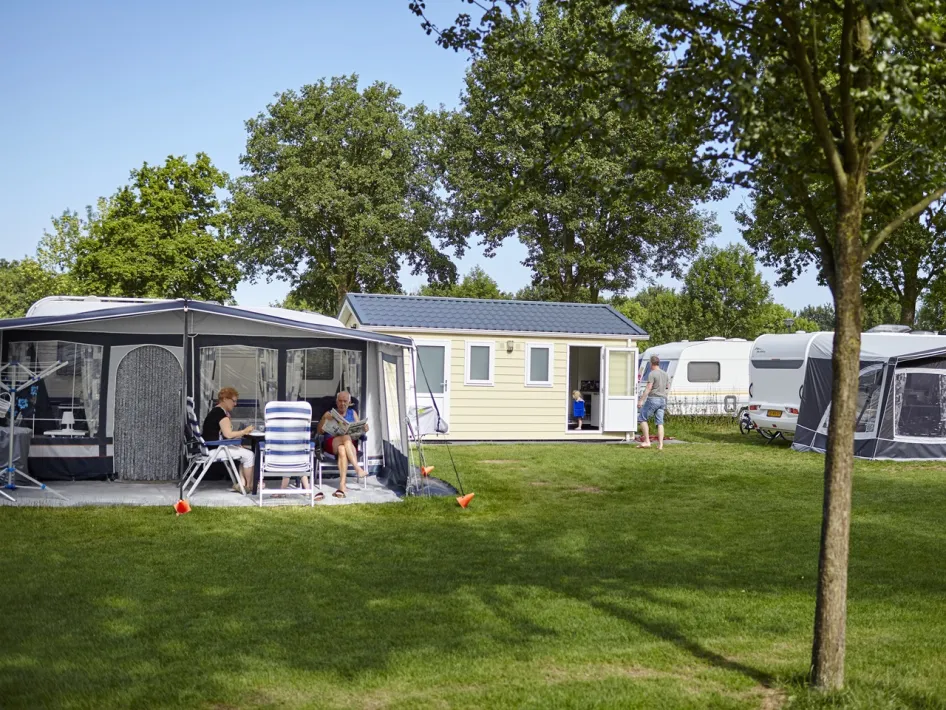 Camper genießen das Draußenleben auf Camping de Leistert, mit Wohnwagen, Vorzelten und einem Chalet auf einem grünen Campingplatz.