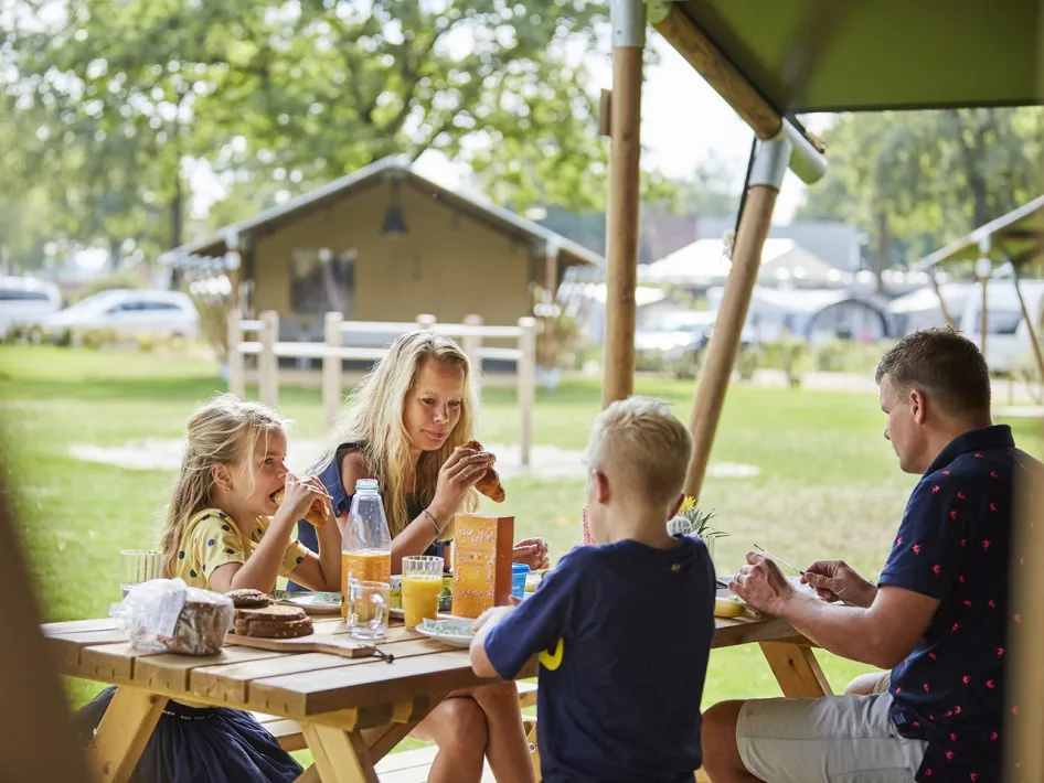 Een gezin ontbijt aan een picknicktafel voor hun glampingtent op camping de Leistert, omringd door zon en groen.