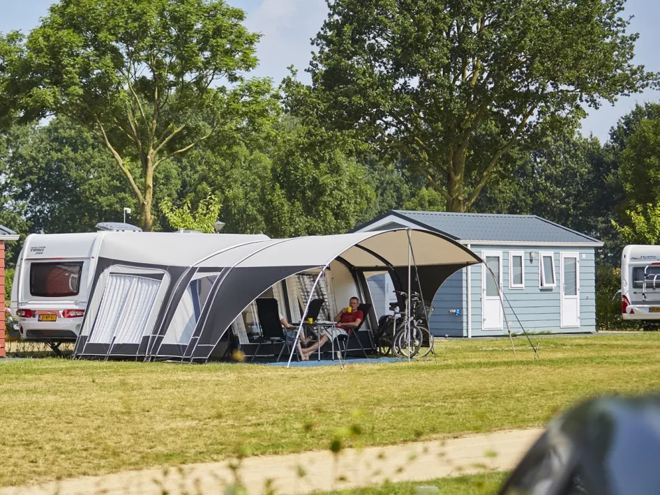 Guests relax by their caravan at Camping de Leistert, under an awning with views over the green grounds.