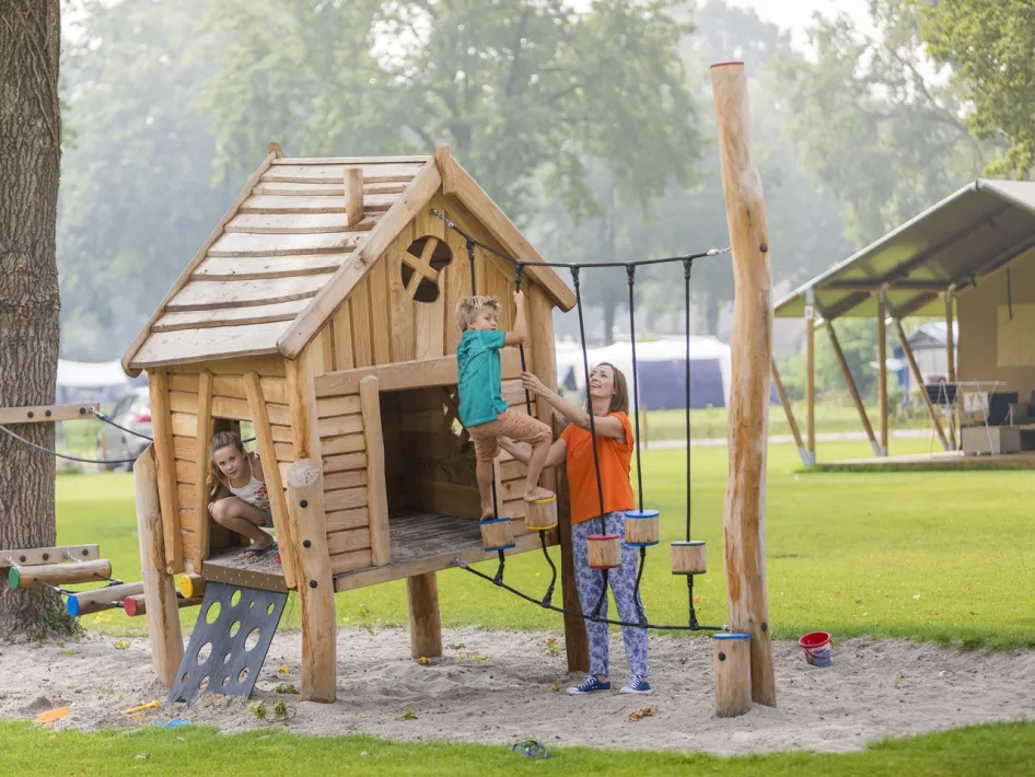 Eine Mutter hilft ihrem Sohn beim Klettern auf einem Holzspielplatz auf Camping de Leistert, im Hintergrund Glamping‑Zelte.