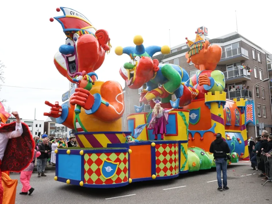 Grote, kleurrijke carnavalswagen met lachende figuren tijdens de carnavalsoptocht, omringd door feestvierders in kostuums.
