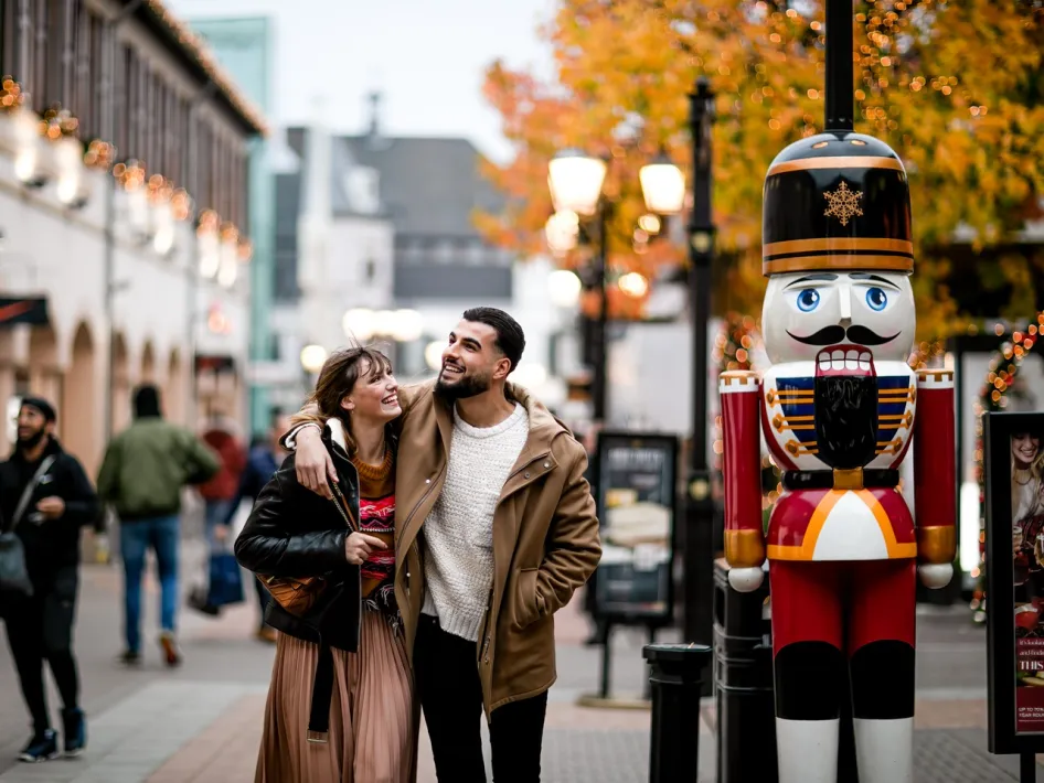 Paar schlendert lachend an einer Nussknackerfigur vorbei während der Christmas Sale im Designer Outlet Roermond, umgeben von festlicher Beleuchtung und Herbstfarben.