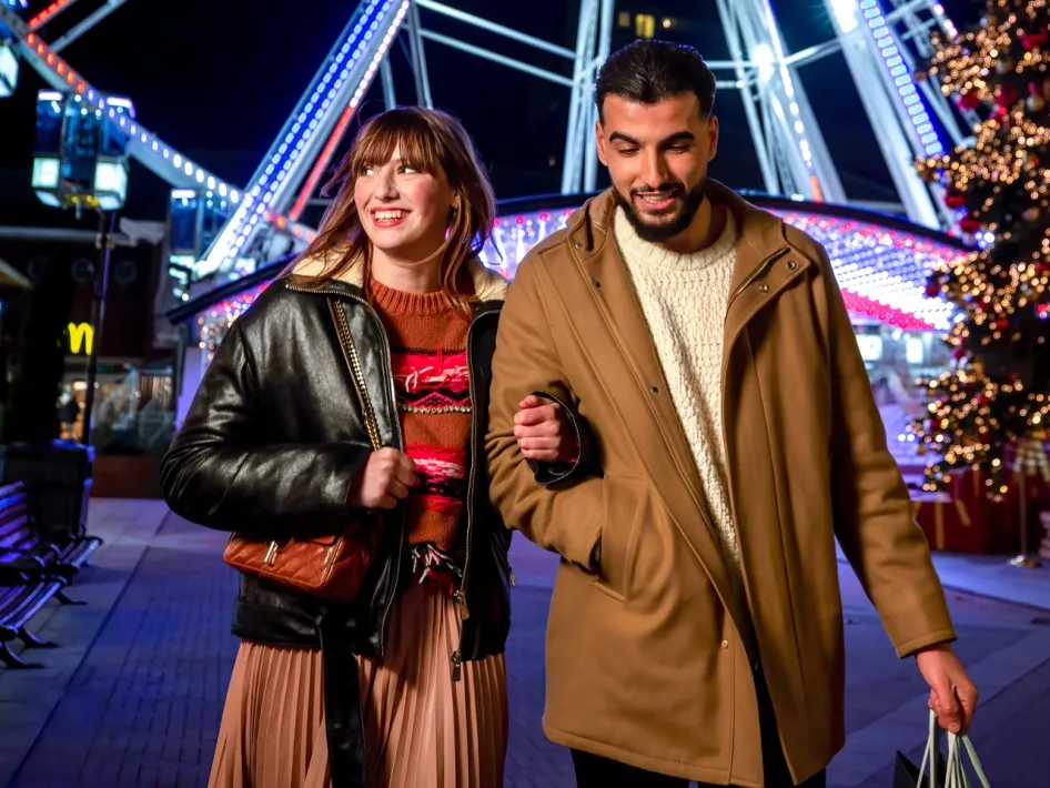 Couple walks smiling past the illuminated Ferris wheel and a Christmas tree during the Christmas Sale at Designer Outlet Roermond.