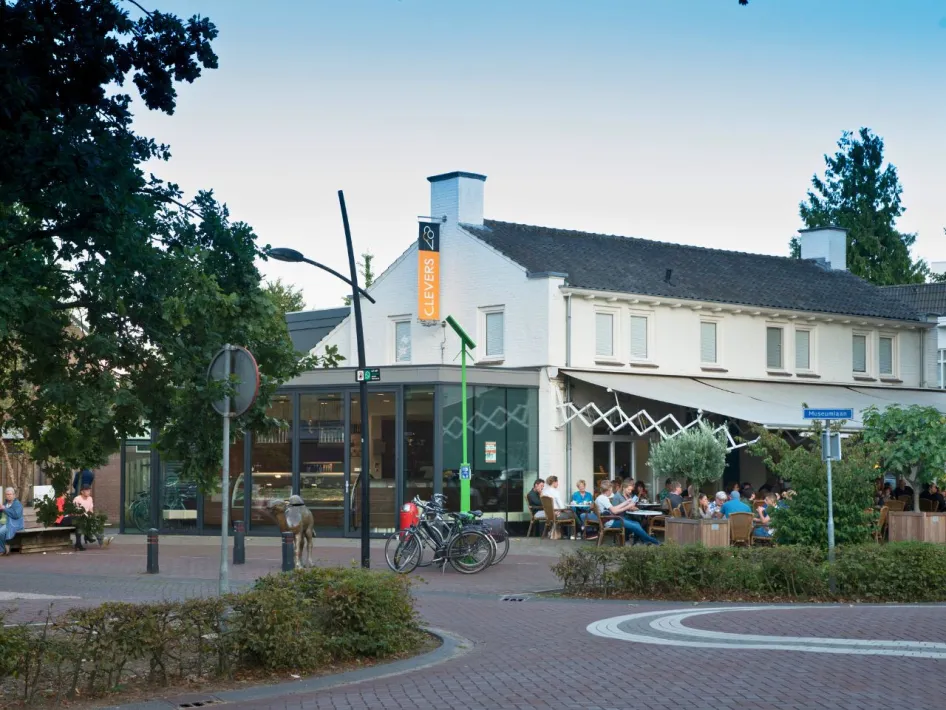 Außenansicht der Eisdiele Clevers Overloon mit einer Terrasse voller Gäste, Fahrrädern und Grünpflanzen auf einem Dorfplatz.