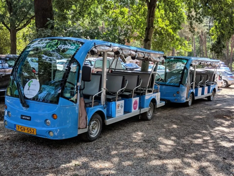 Twee blauwe elektrische treintjes van de Geluksexpres staan geparkeerd onder bomen op een onverharde parkeerplaats in Roerdalen.