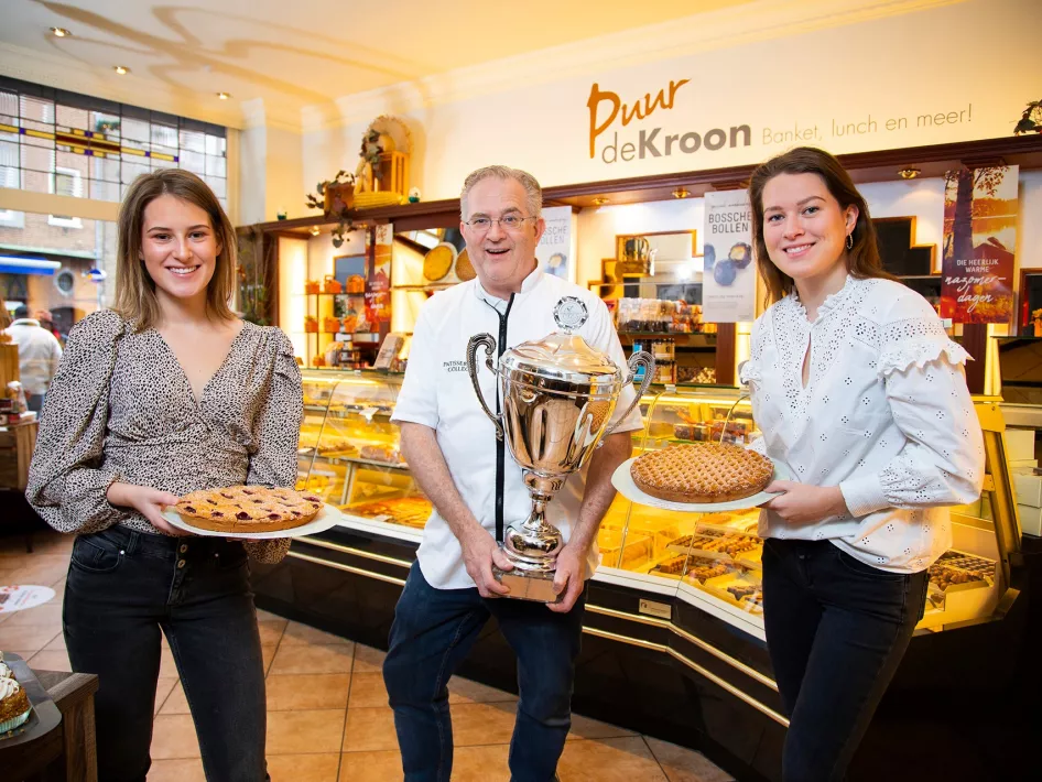 Auf diesem Foto präsentieren drei Personen stolz verschiedene Backwaren in einer Konditorei. Eine Person hält einen großen Pokal, was auf preisgekröntes Gebäck hinweist. Die Umgebung zeigt eine warme, einladende Atmosphäre mit Vitrinen voller köstlicher Leckereien. Das Schild im Hintergrund zeigt den Namen „Puur de Kroon“ und betont den Schwerpunkt auf Konditorei und Mittagstisch.