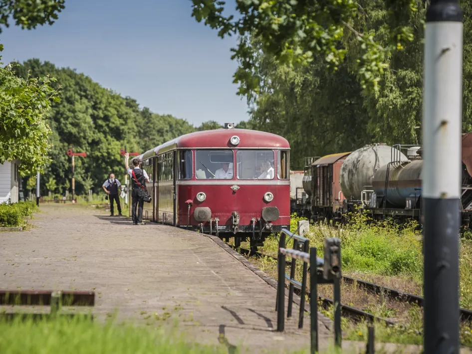 Nostalgische rode railbus arriveert op station tussen het groen, begeleid door treinpersoneel.