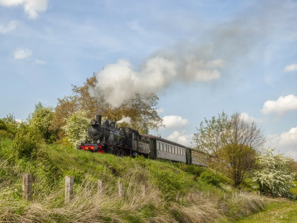 Stoomtrein rijdt door glooiend landschap met rookpluim onder blauwe lucht.