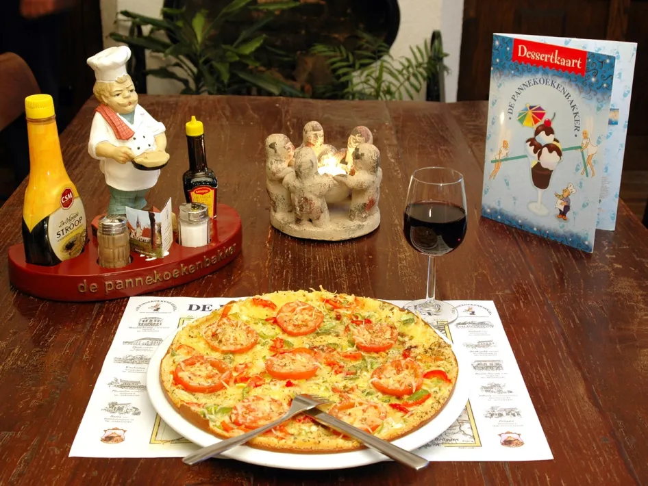 Herzhafter Pfannkuchen mit Gemüse, serviert im De Pannekoekenbakker, mit Sirup, Dessertkarte und einem Glas Wein auf dem Tisch.