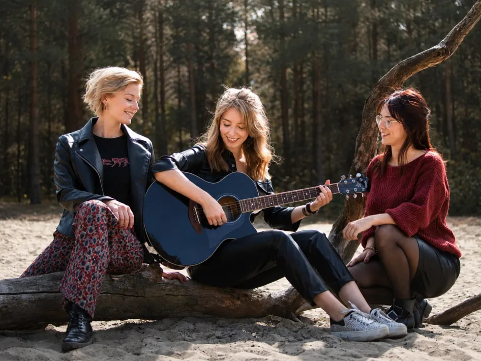 Drie leden van zangtrio JEM zitten buiten bij elkaar, met een akoestische gitaar.