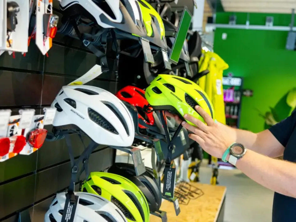A person looks at a bright yellow bicycle helmet in a bicycle store. In the background, several helmets in various colors hang from a display, along with bicycle lights and other accessories.