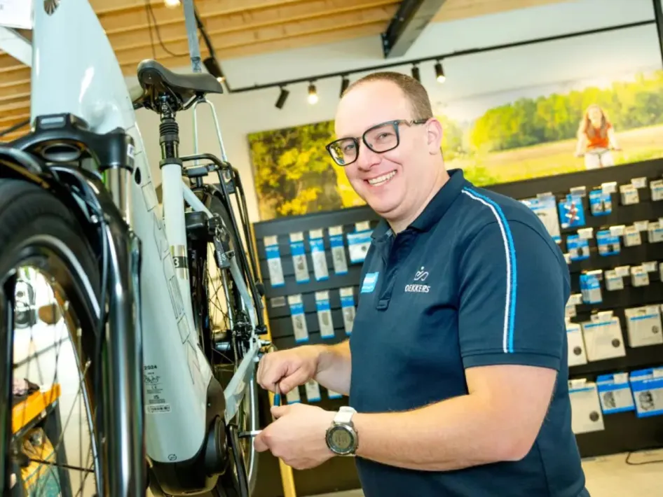 Een vriendelijke fietsenmaker in een blauw poloshirt van Dekkers Tweewielers repareert een witte e-bike in de werkplaats van de fietsenwinkel. Op de achtergrond hangen fietsaccessoires aan de muur.