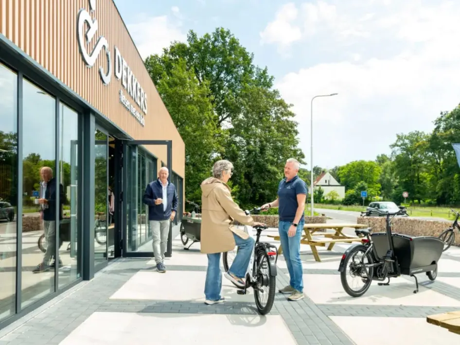 A modern bicycle store called Dekkers Tweewielers with large glass windows and a wooden facade. In front of the store are people engaged in conversation and trying out an e-bike. A cargo bike and a picnic table are visible in the background.