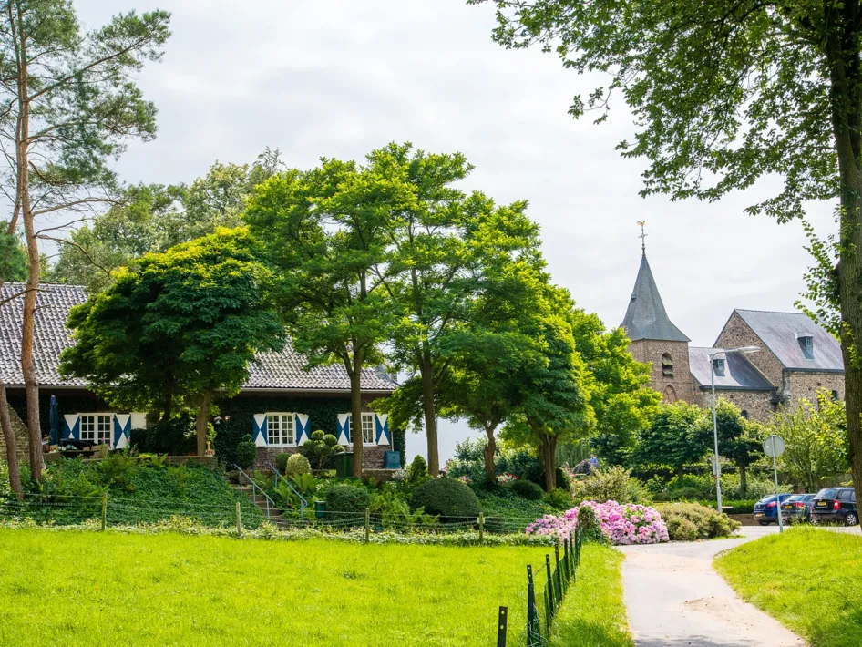 A green landscape with a half-timbered house in the foreground and an old church in the background, surrounded by trees and blooming shrubs.