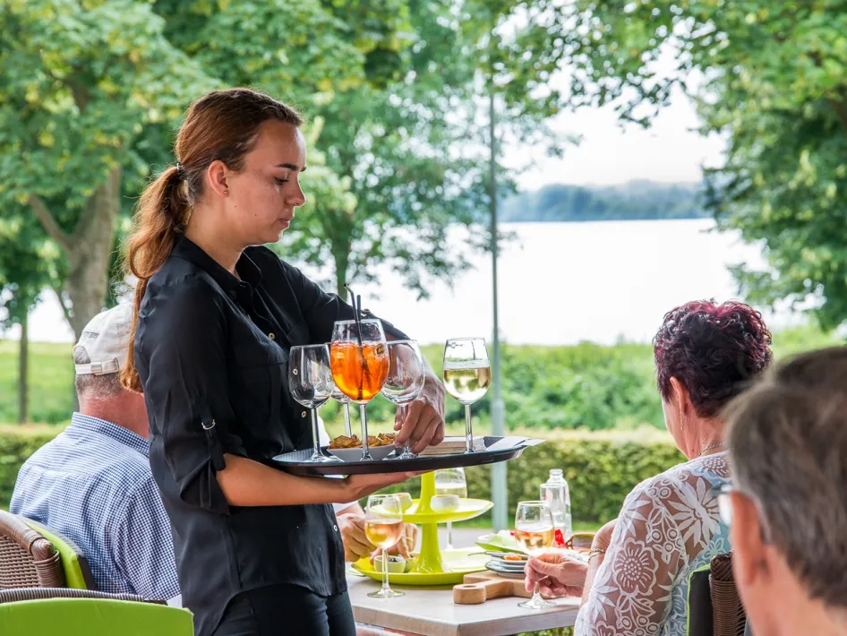 Een serveerster zet drankjes op tafel bij een groep mensen op een terras met uitzicht op het water, omringd door bomen in zomerse sfeer.
