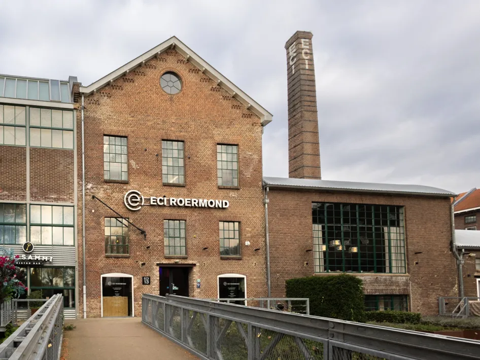 Außenansicht der ehemaligen Fabrik des ECI Roermond mit Backsteinfassade, hohem Schornstein und einem Steg zum Eingang.