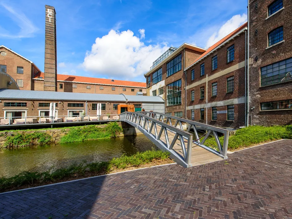 Buitenzijde van ECI Roermond met industrieel erfgoed, schoorsteen en brug over het water.