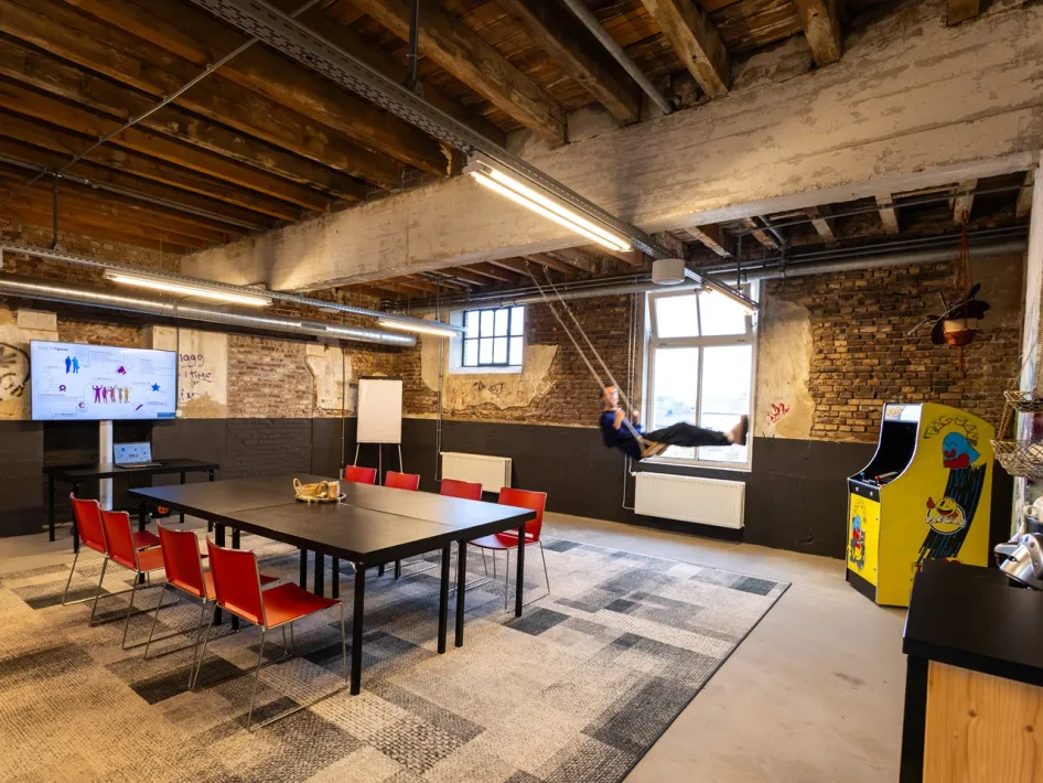 Industrieller Besprechungsraum im ECI Roermond mit einem großen schwarzen Tisch, roten Stühlen, einer Schaukel und einem gelben Pac-Man-Spielautomaten.