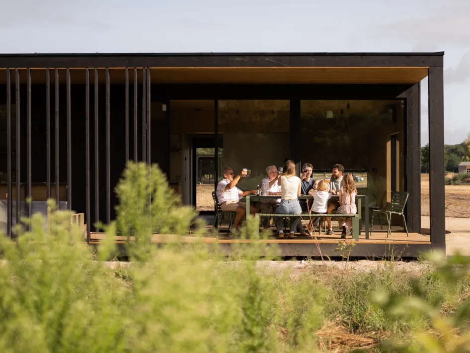 Familie zit samen aan een grote tafel op het overdekte terras van een lodge van Eco by Nature in Heythuysen, omringd door groen.
