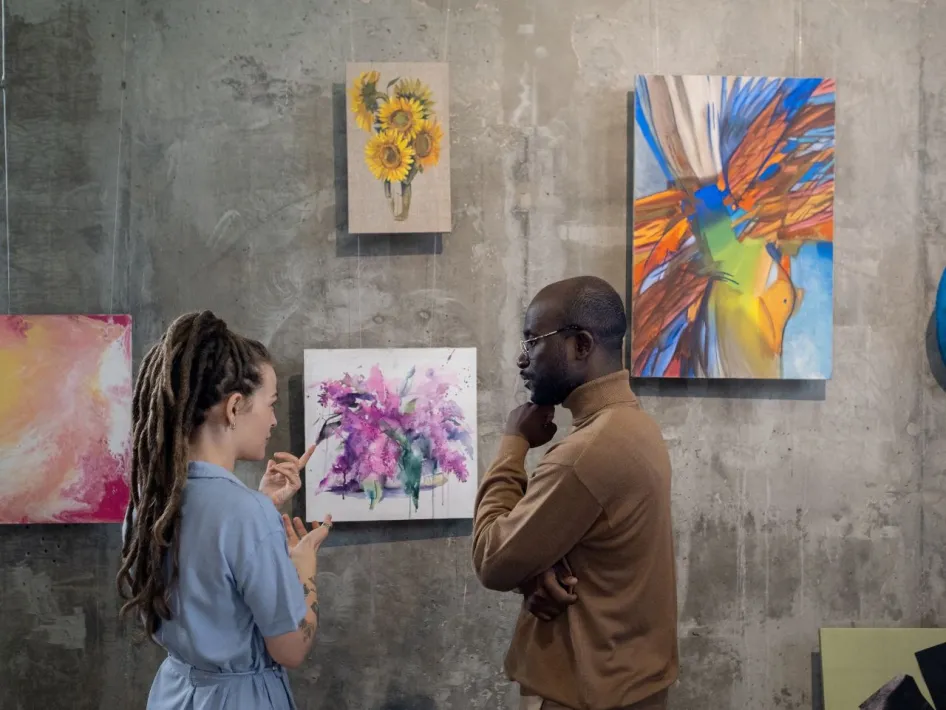 Twee personen staan voor een betonnen muur en bekijken samen kleurrijke schilderijen tijdens een kunstexpositie; één persoon wijst naar een abstract werk terwijl de ander geïnteresseerd luistert.