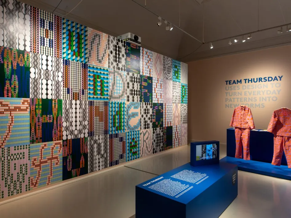 Overzicht van een wand met grafische patronen, een blauw informatief meubel en kledingstukken in roze-oranje design, onderdeel van een designexpositie.