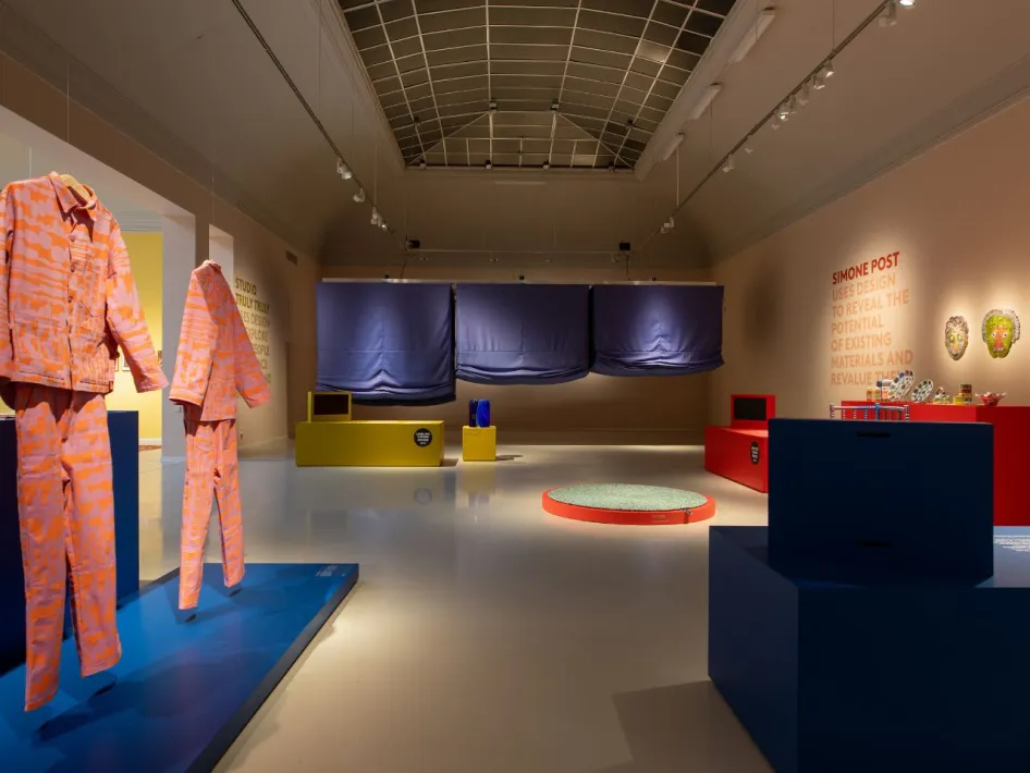Tentoonstellingsruimte met kleurrijke installaties en designobjecten, waaronder roze-oranje kledingstukken, blauwe doeken en informatieve displays.