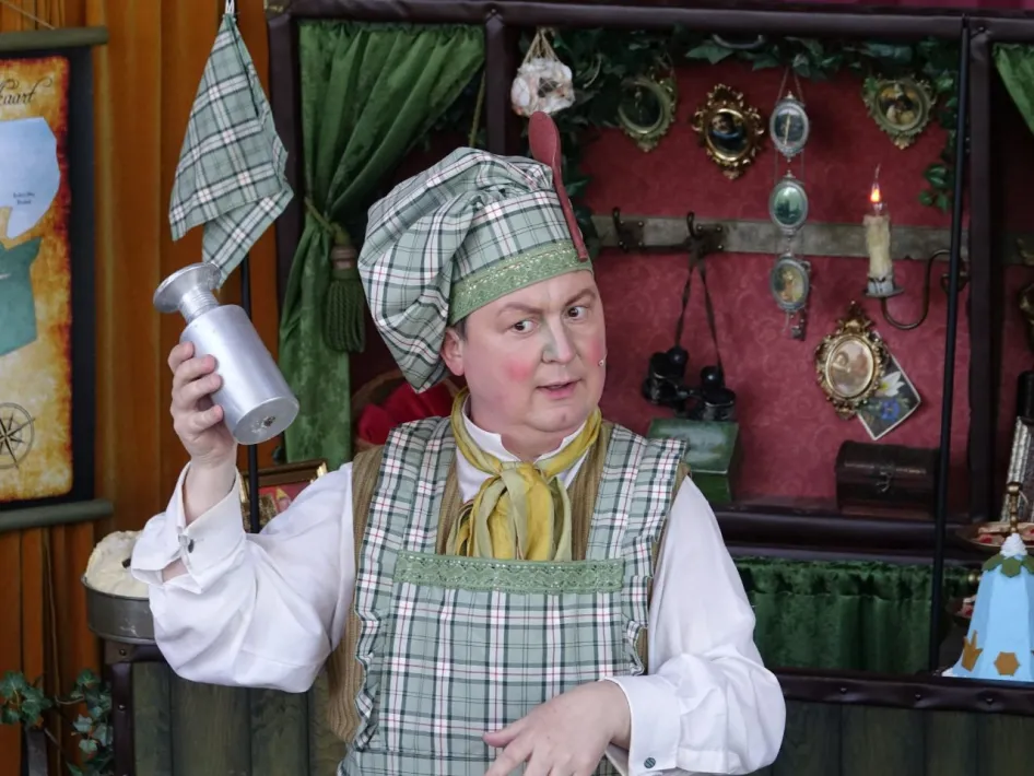 Acteur in geruite kostuumschort en hoofddeksel houdt een zilverkleurige beker omhoog tijdens een voorstelling, met een theatraal ingerichte achtergrond vol accessoires.