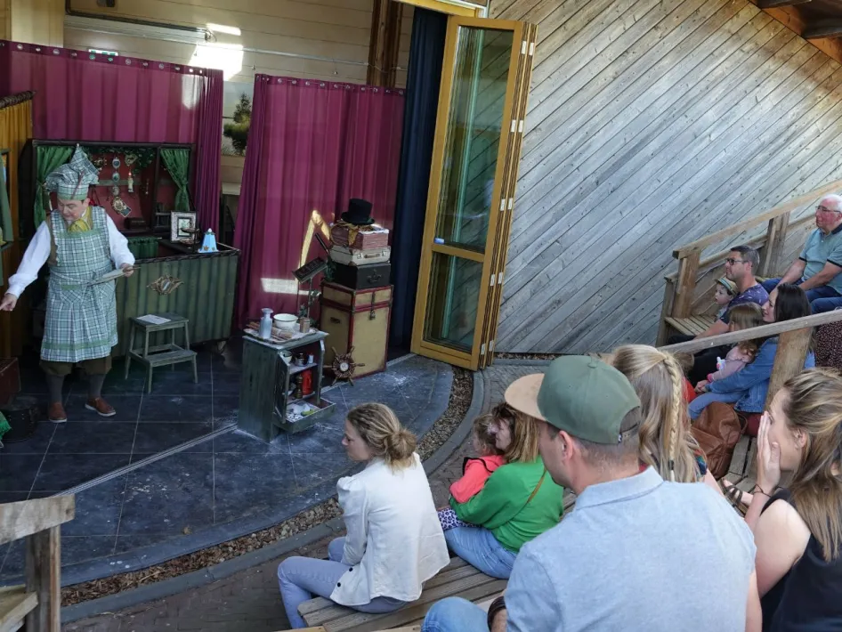 Openluchttheater met houten tribunes waar families en kinderen aandachtig kijken naar een theatervoorstelling met kleurrijk decor en een verklede speler.