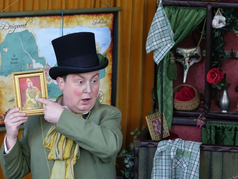Acteur in kostuum en hoge hoed toont een ingelijste foto tijdens een theatervoorstelling, met op de achtergrond een kaart en decoratieve elementen.