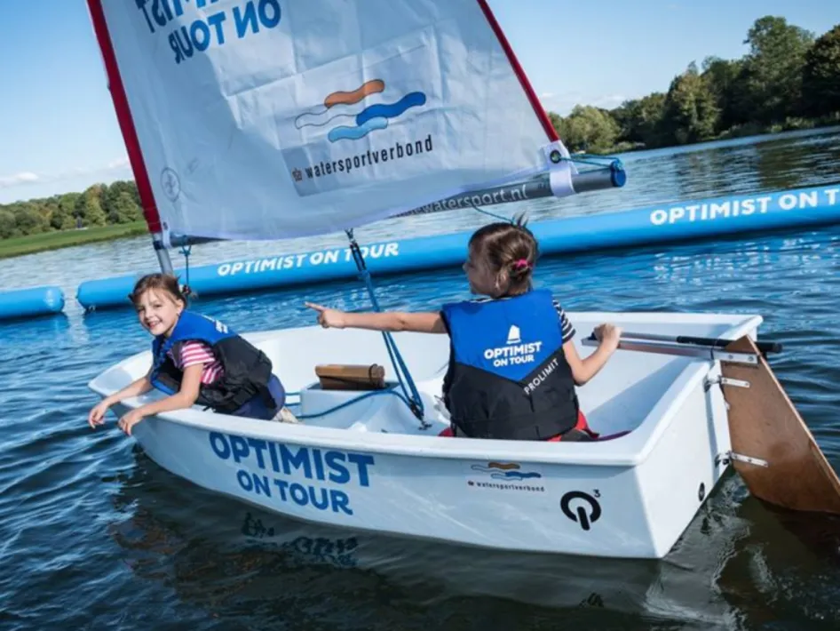 Twee kinderen in een klein zeilbootje met de tekst "Optimist on Tour" zeilen op rustig water, omringd door natuur.