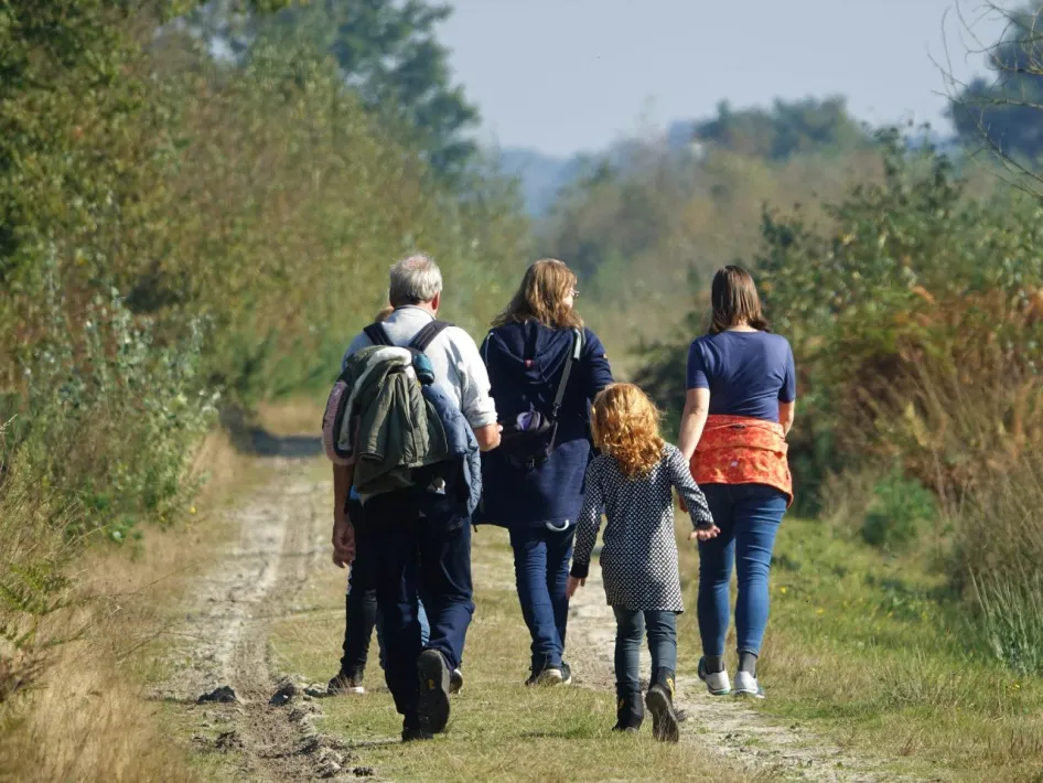 Een groep mensen, waaronder een kind, geniet van een ontspannen wandeling over een zandpad in een groen natuurgebied op een zonnige dag.