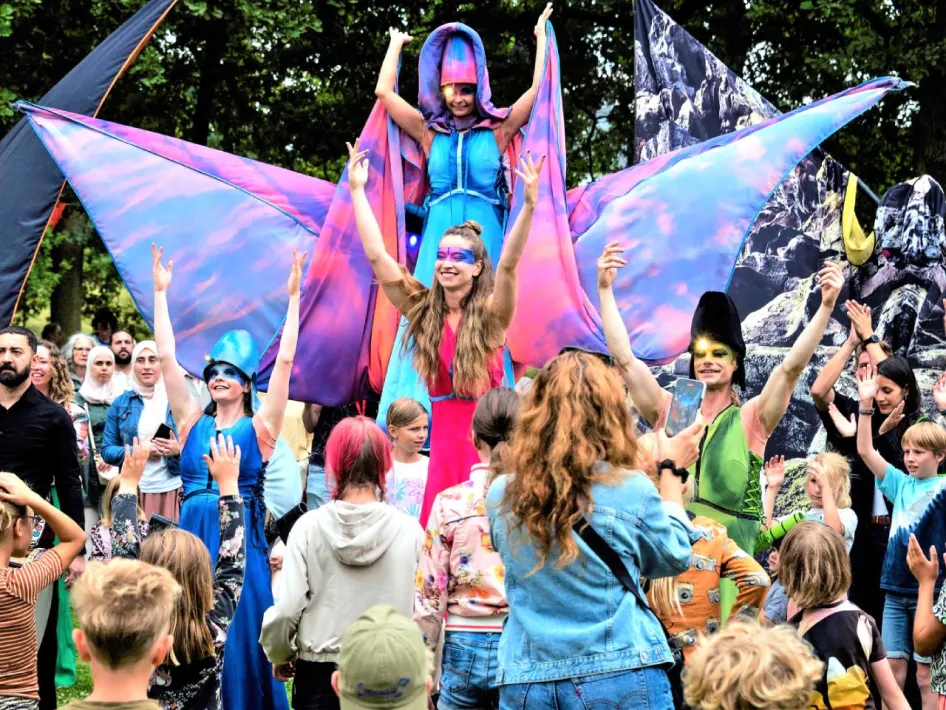 Kinderen en artiesten genieten van kleurrijk straattheater tijdens De Gekleurde Stad Roermond, met acrobaten, opvallende kostuums en publiek op een plein in het centrum.