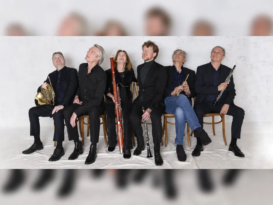 Zes musici van het Hexagon Ensemble zitten naast elkaar met hun blaasinstrumenten tijdens een groepsfoto.