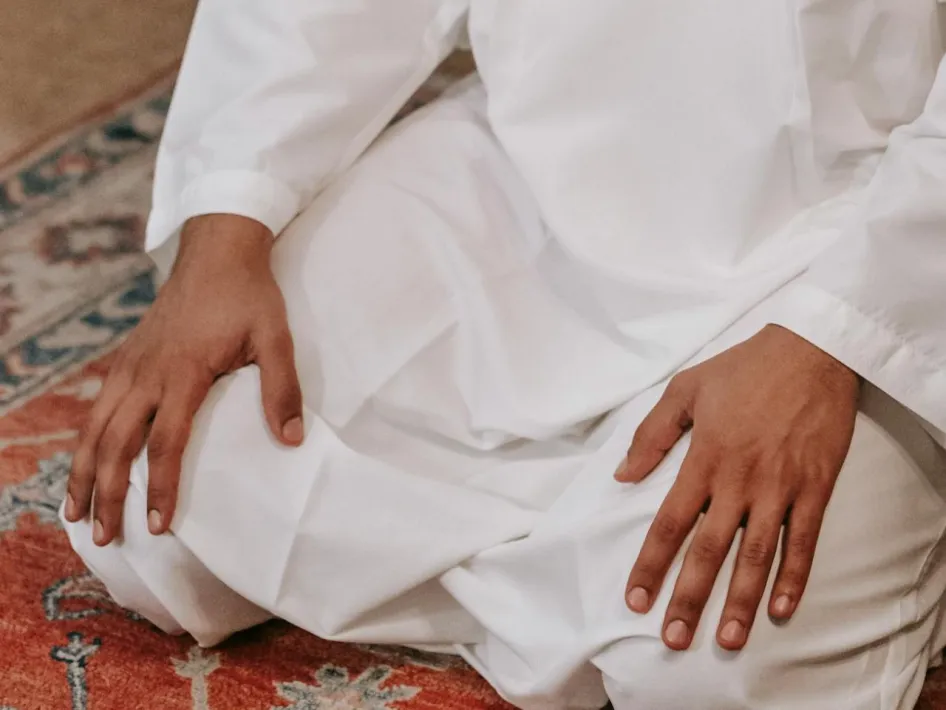 Meditatie in witte kleding, zittend in kleermakerszit met handen op de knieën op een kleurrijk tapijt.