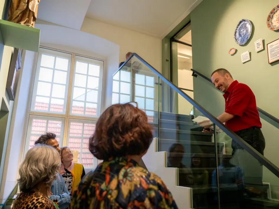 Tijdens een instaprondleiding in Museum W vertelt een gids in een rood shirt op de trap aan een kleine groep bezoekers over de collectie in een lichte museumruimte met groene muren en grote ramen.