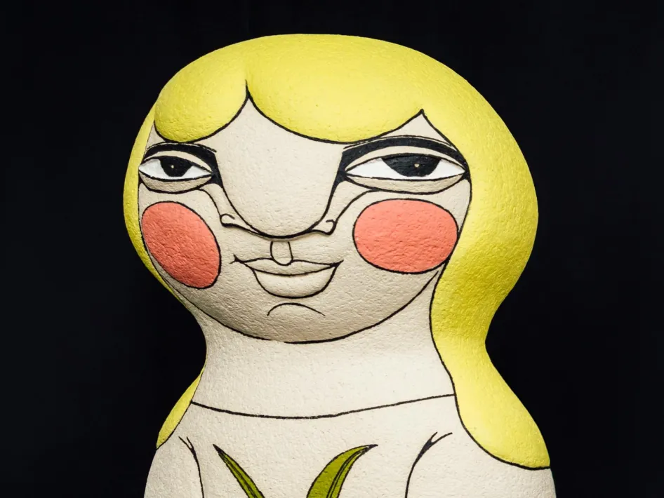 Keramische Skulptur einer weiblichen Figur mit gelbem Haar, rosigen Wangen und einem stillen Blick vor dunklem Hintergrund.