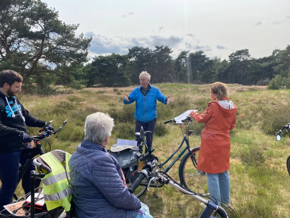 Stadsgids geeft uitleg aan deelnemers van de Fietstocht Rondje Weert tijdens een stop op de Boshoverheide, met fietsen en heide op de voorgrond.