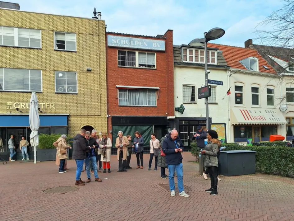 Deelnemers aan een stadswandeling staan op een plein in het centrum van Weert en luisteren naar de gids.