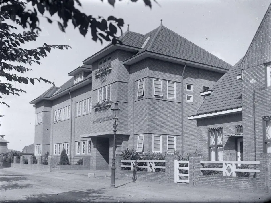 Historische zwart-witfoto van een voormalig schoolgebouw in Weert met bakstenen gevel en klassieke straatlantaarn.