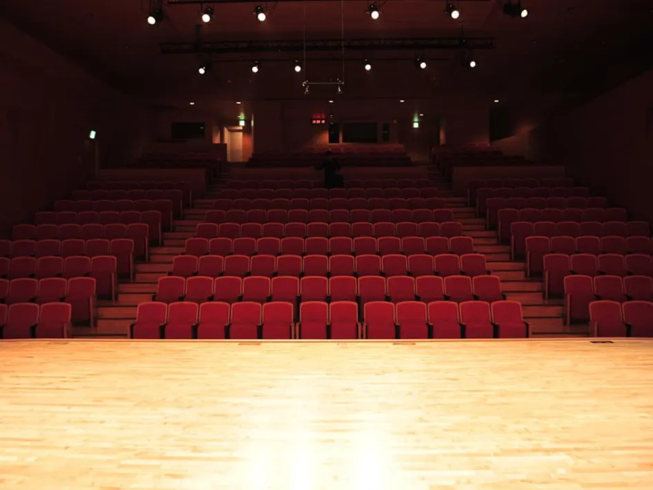 Lege theaterzaal met rode stoelen en houten podium, gezien vanaf het toneel richting de tribune met sfeerverlichting bovenin.