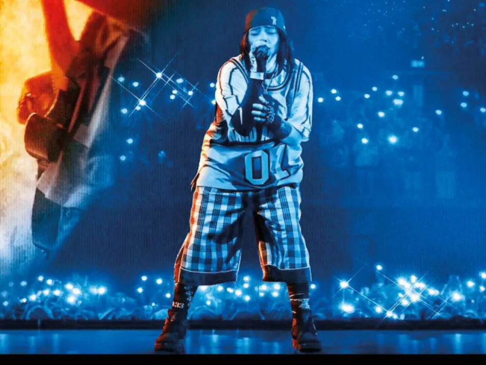 Concertfilm Billie Eilish: Billie Eilish staat zingend op het podium in blauw licht, met een microfoon in haar hand en een verlichte zaal vol publiek op de achtergrond.